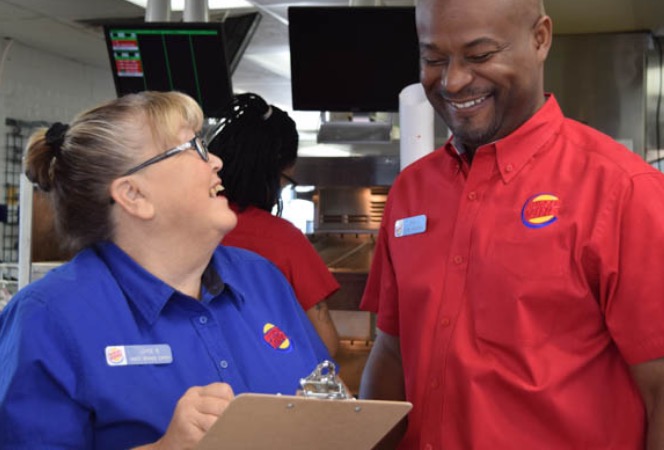 A female Burger King employee holding a clipboard laughing with a male Burger King employee while they consult her clipboard notes