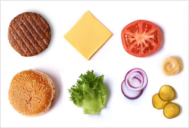 An overhead view of a hamburger spread out with numerous condiments on a table: a cooked hamburger patty, a cheese slice, a slice of tomato, a dollop of mustard, sliced pickles, a few raw onion rings, a leaf of lettuce and a sesame seed bun