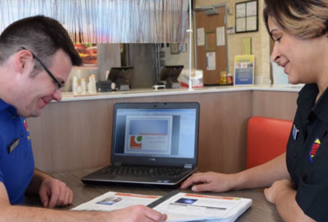 Two Burger King employees, one male and one female, sitting at a table with a laptop computer and going through hard-copy notes