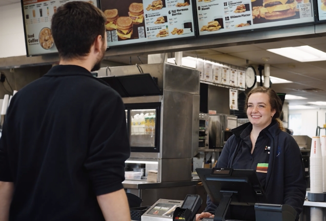 A woman working at a Burger King checkout interacts with a male customer