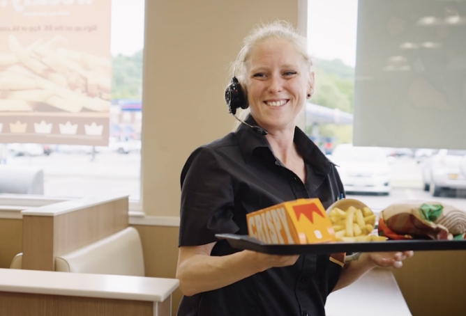 A female Burger King employee, in a headset, smiling while delivering a tray full of food