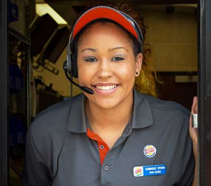 A female Burger King employee, in a headset, smiling while working at a drive-through window